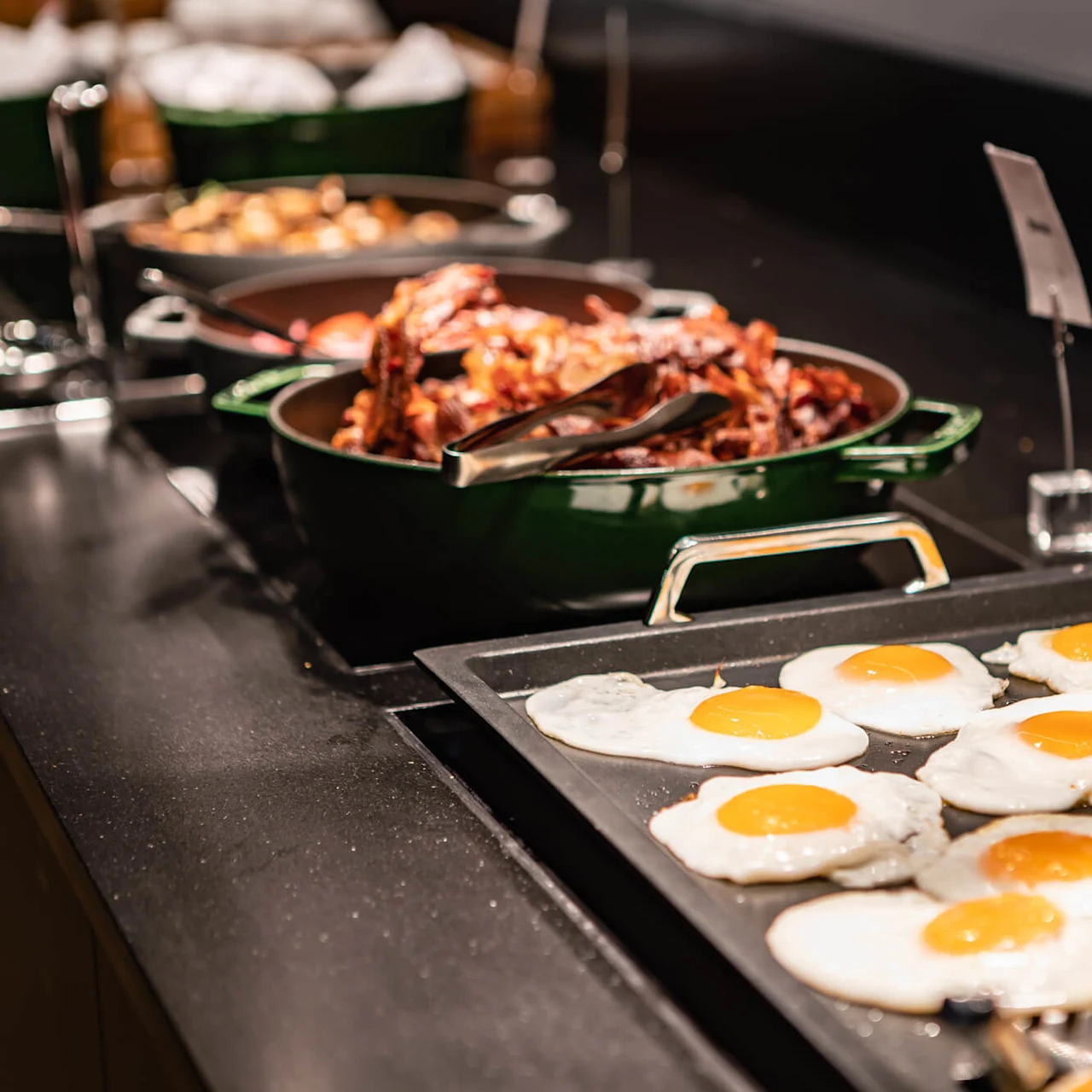 Fried eggs sit on a tray with other breakfast dishes like bacon and potatoes in the background on a buffet counter.