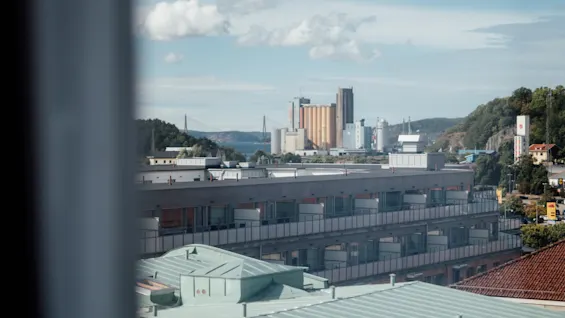 Byggnader med gröna tak ses genom ett fönster, med en avlägsen industriell silhuett och kullar under en delvis molnig blå himmel.