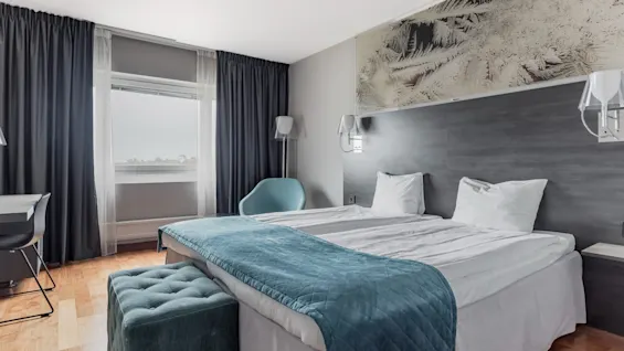 A modern hotel room with two beds, a desk, and a window, featuring a blue throw blanket and dark curtains.