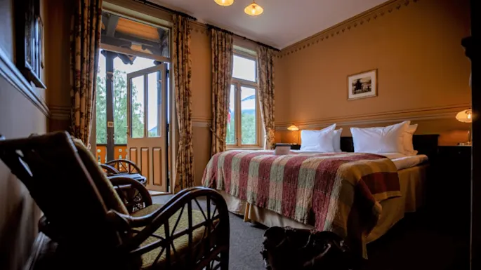 A cozy hotel room features a double bed with a patterned quilt and pillows, an open glass door leading to a balcony, and wicker chairs in a warmly lit setting.