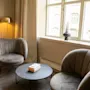 To grå lenestoler vender mot et rundt svart kaffebord som holder en bok, i et mykt opplyst rom med et vindu, beige gardiner, en gyllen lampe og potteplante.