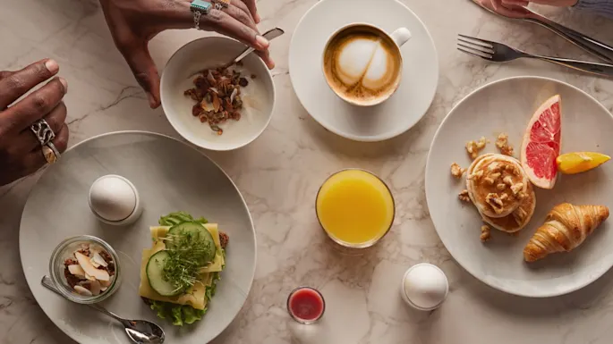 Et varieret morgenbord: en person rører i yoghurt med granola, en anden holder bestik. Tallerkener med æg, grønt, ristet brød, grapefrugt, en croissant; cappuccino, juice, og syltetøj på marmorbord.