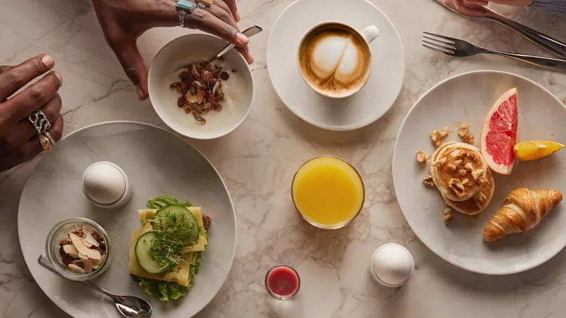 A diverse breakfast spread: one person stirs yogurt with granola, while another holds cutlery. Plates include eggs, greens, toast, grapefruit, a croissant; cappuccino, juice, and jam on marble table.