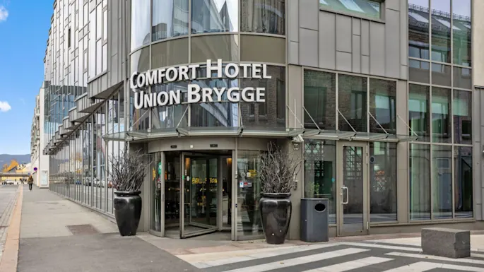 A modern hotel entrance features large glass doors beneath the sign "COMFORT HOTEL UNION BRYGGE." Two tall planters flank the entrance in an urban setting with a crosswalk nearby.