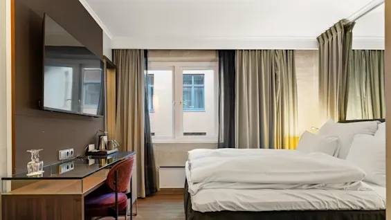En velopredt seng med hvitt sengetøj, står ved siden av et skrivebord med stol, TV, kedel og glas. Høje vinduer med gardiner giver naturligt lys.