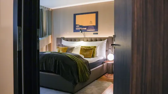 Et koselig hotellrom med en stor seng, grønne og grå sengetøy, og en moderne lampe, sett gjennom en åpen døråpning.
