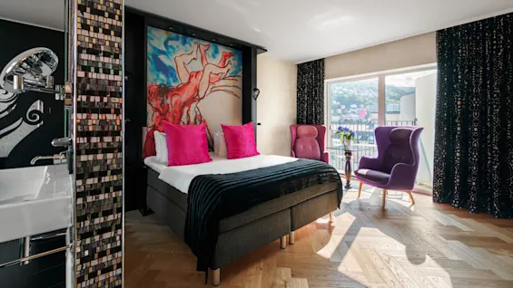 A modern bedroom features a bed with pink pillows, vibrant artwork on the wall, and two purple chairs by a large window overlooking a cityscape.