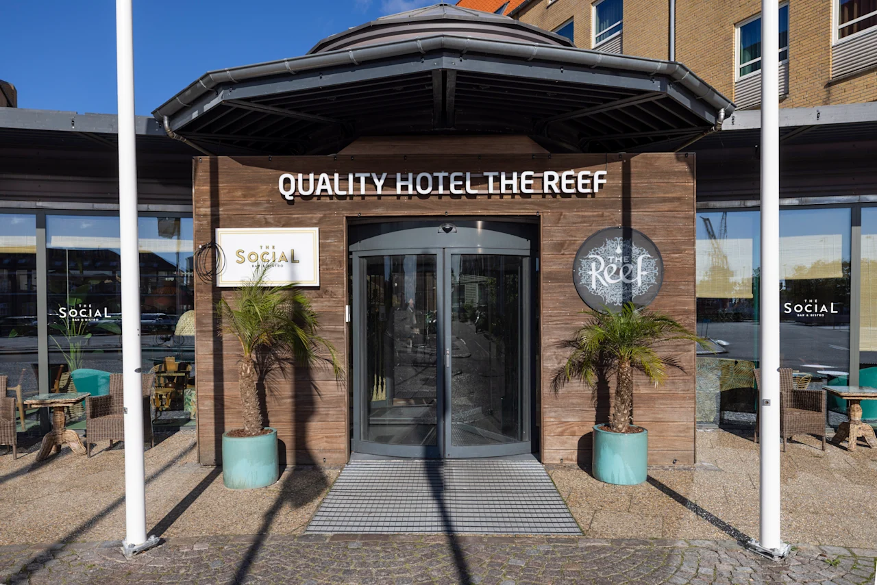 En ingång av glas märkt "QUALITY HOTEL THE REEF" med sido-skyltar "SociAL" och "Reef"; omgiven av växter i krukor på en skuggig uteplats med sittplatser utomhus.