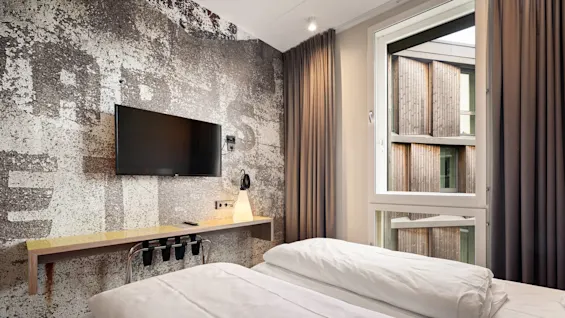 A wall-mounted TV hangs in a modern bedroom with a textured wall, beside a large window showing wooden buildings outside. A shelf below holds a lamp and remote.