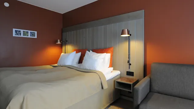 Bed covered with beige bedding and white pillows sits against an orange wall, flanked by two wall lamps and a small nightstand. A gray sofa is nearby. Three small pictures adorn the wall.