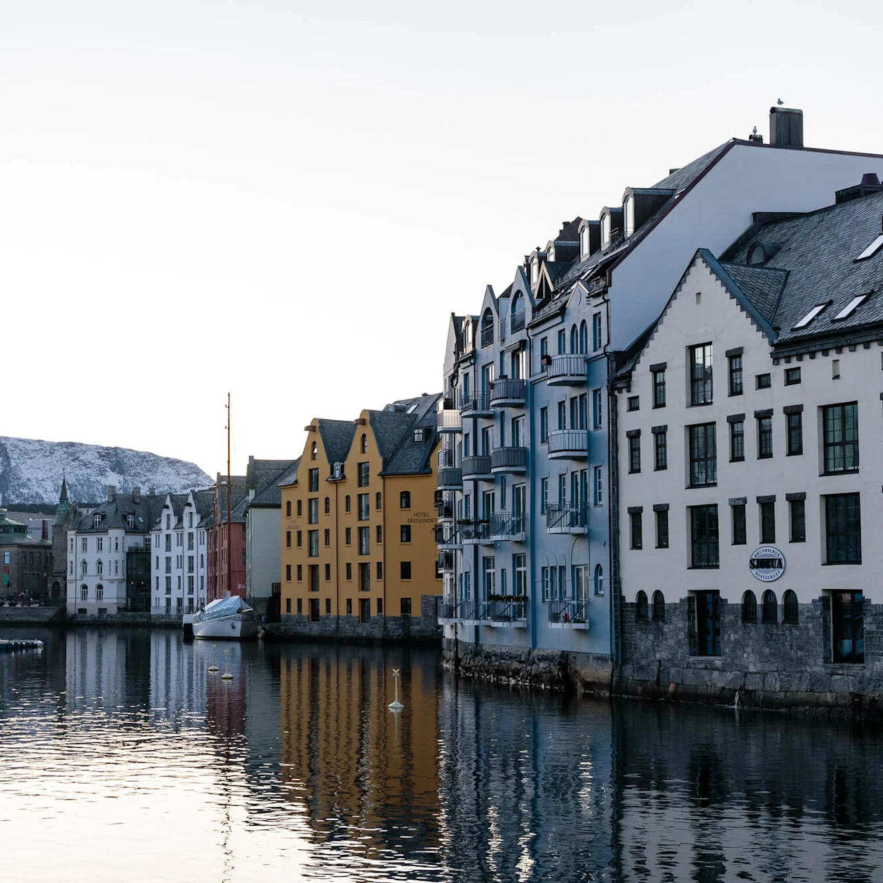 Colorful boats dock quietly beside canal-front buildings; snowy mountains rise in the background. Two signs read "Hotel Brosundet" and "Apotekergata" on the buildings.