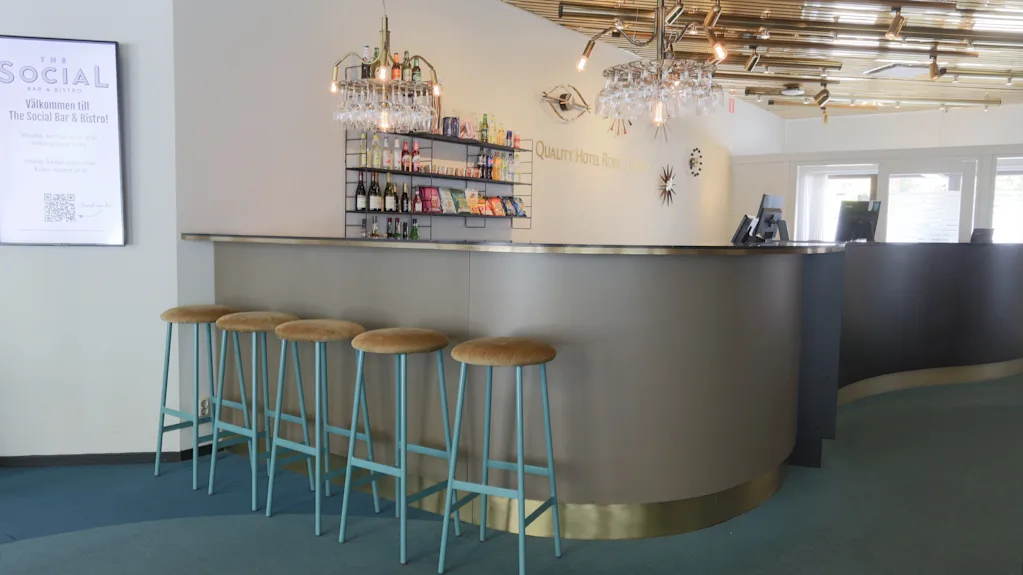 Bardisk med fem krakker, belyste hyller viser flasker og snacks. Krystallkroner henger over. Hvit veggtekst: "Quality Hotel Sarpsborg". Skilt: "SOCIAL Welcome to The Social Bar & Bistro".