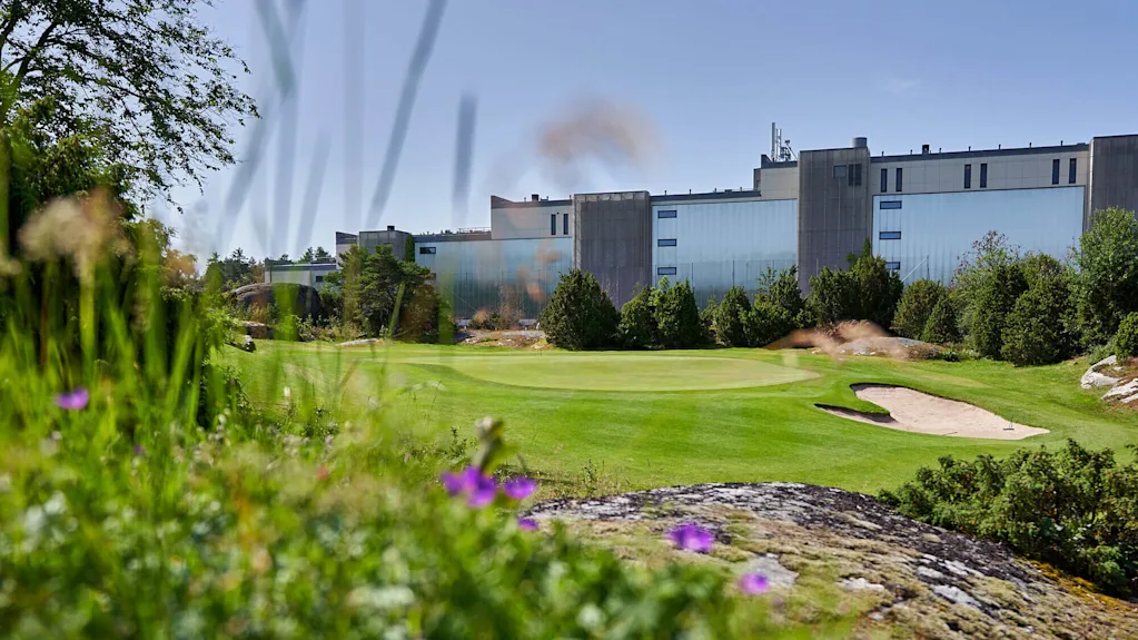 Grass surrounds a golf course with a sand trap. In the background, a large industrial building stands amidst trees under a clear sky.