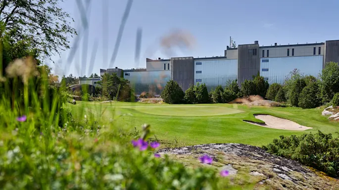 Gress omgir en golfbane med en sandbunker. I bakgrunnen står en stor industribygning blant trær under en klar himmel.