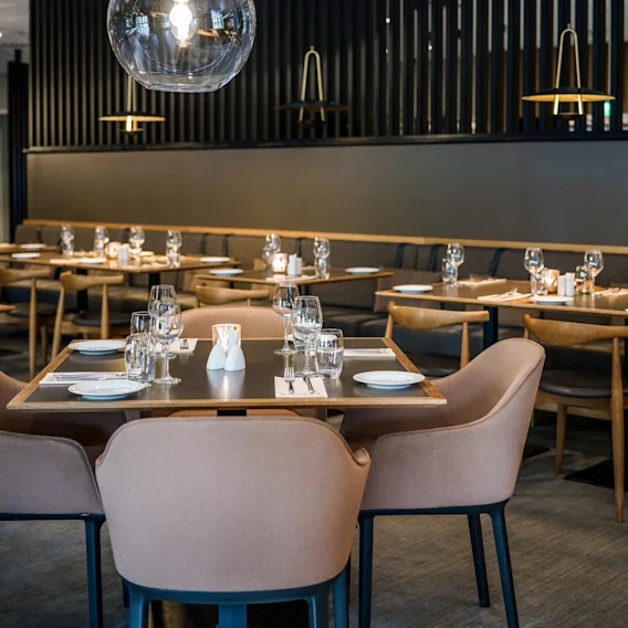 Et dekket bord med elegant glass og servietter, omgitt av moderne stoler, er plassert i en stilfull, svakt opplyst restaurant med tretrekk og varm belysning.
