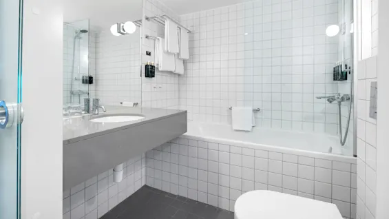 Baderom med hvit flislagt vegg, stort speil, grått benkeplatevask og badekar med dusj. Hyller holder håndklær og toalettsaker og skaper en ren, moderne stil.