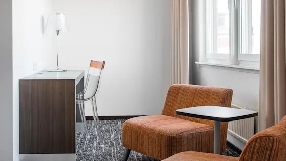 Ett mysigt rum med ett skrivbord, genomskinlig stol och lampa, bredvid orange klädda stolar och ett litet bord vid ett fönster med beige gardiner.