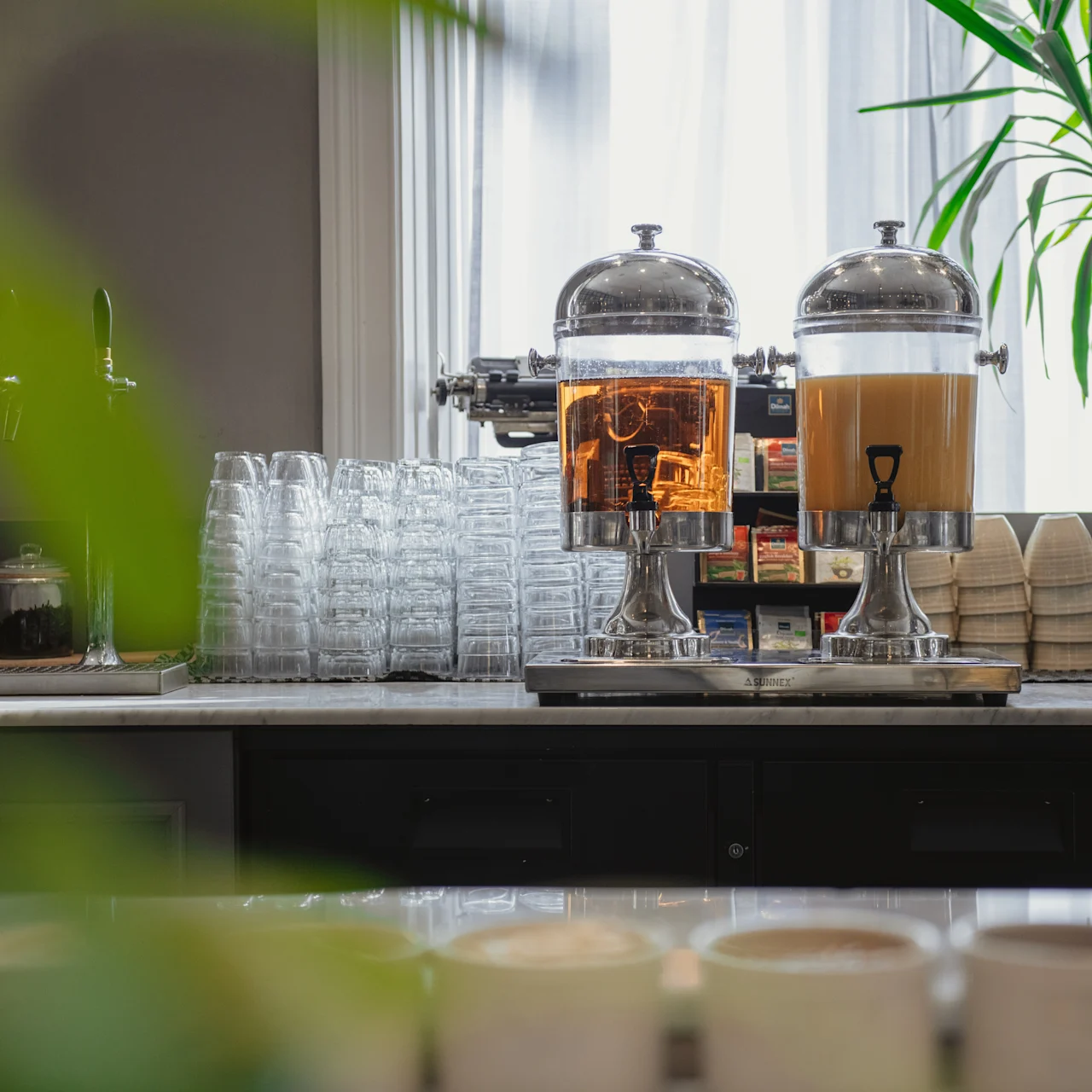 Två dryckesdispenserar, en med kaffe och den andra med mjölk, står på en marmorbänk omgivna av staplade glas och koppar. En växt och naturligt ljus dekorerar utrymmet.