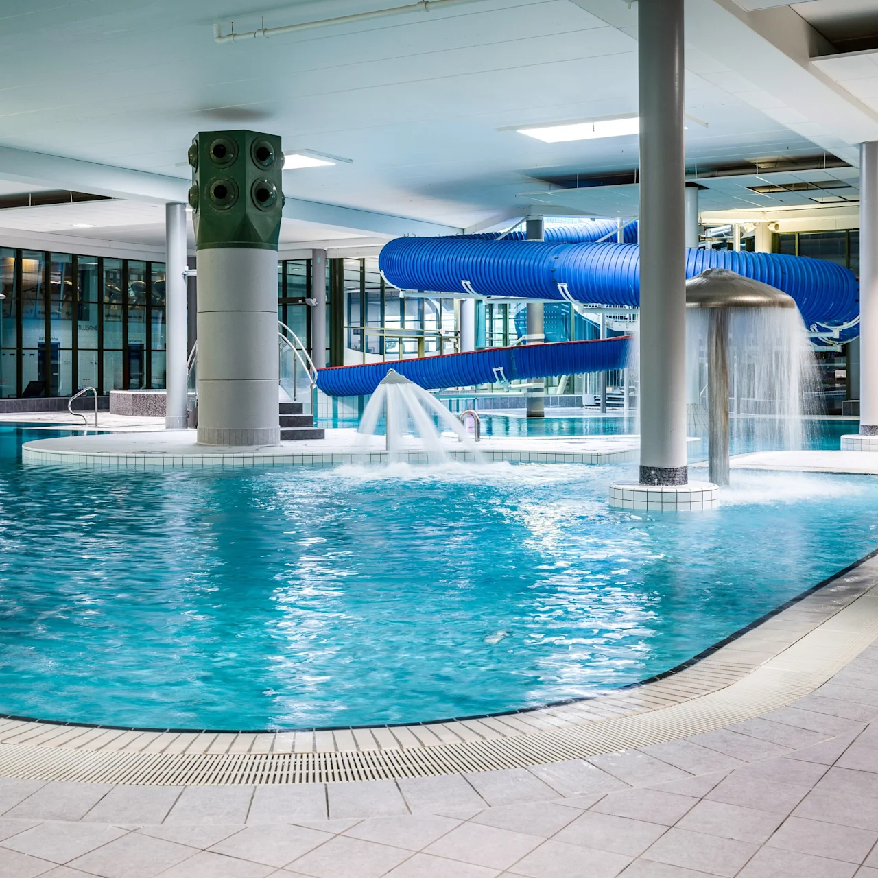 Indoor pool with blue water slide and water fountain; surrounded by tiled flooring and large windows. No text present.