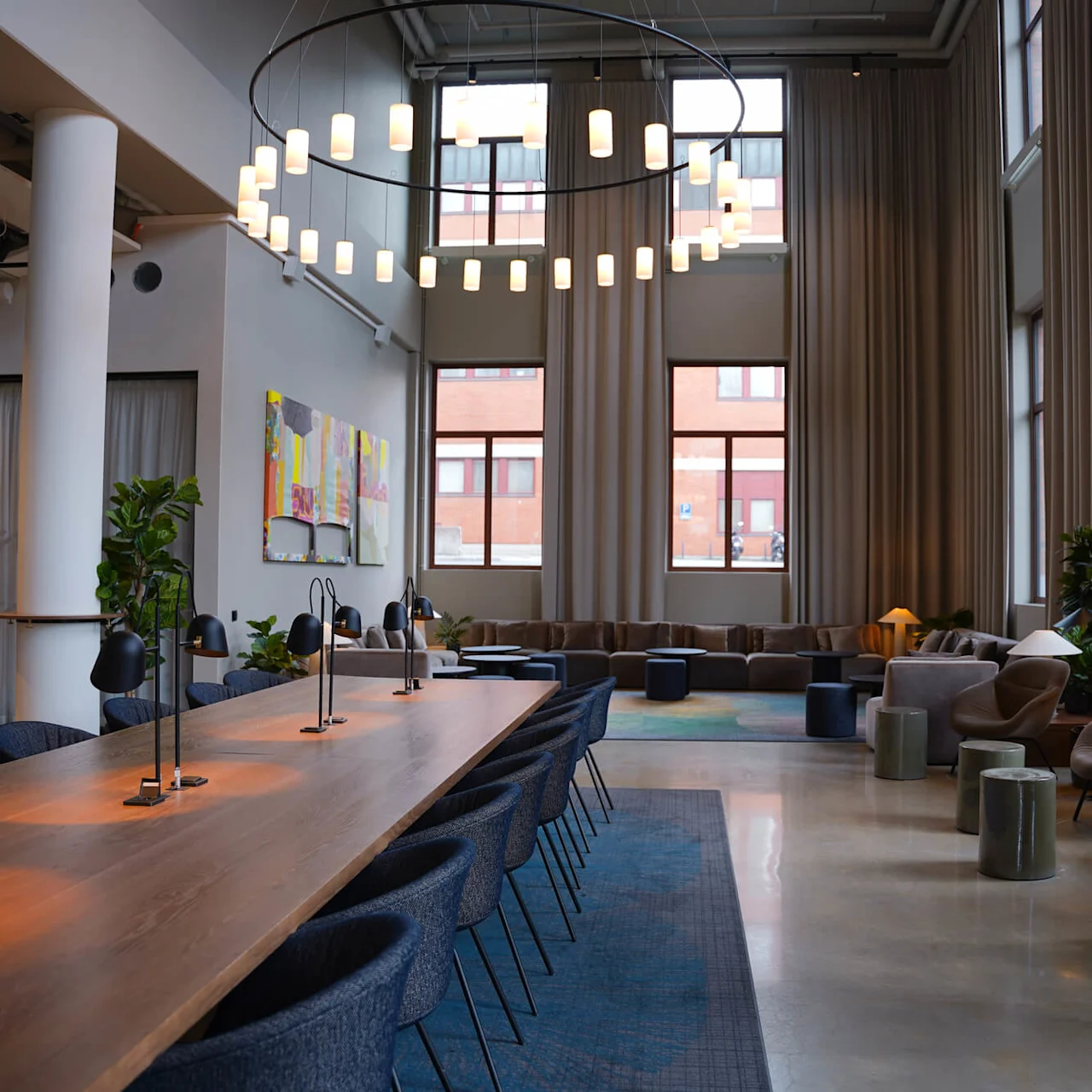 A modern office lounge area with sofas, a long wooden table with chairs, and hanging lights against large windows.