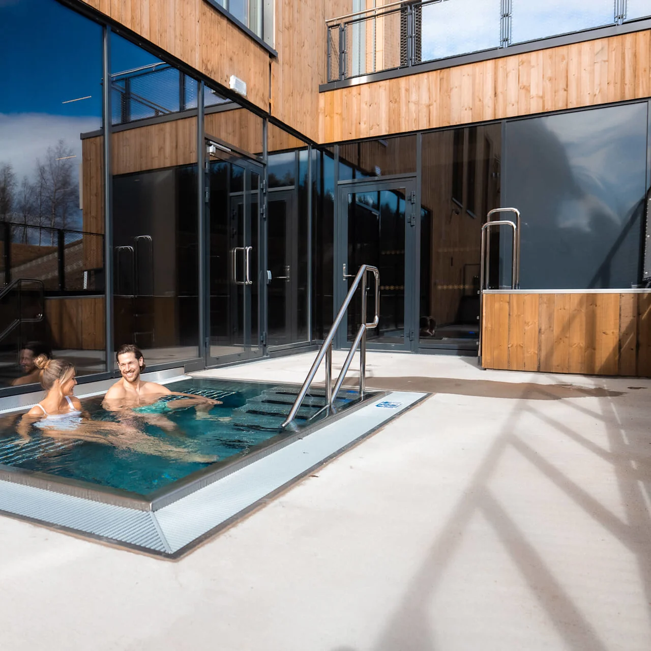 Två personer kopplar av i ett utomhusbubbelpool bredvid en modern träbyggnad under en klarblå himmel på Lily Country Club i norska Klöfta.