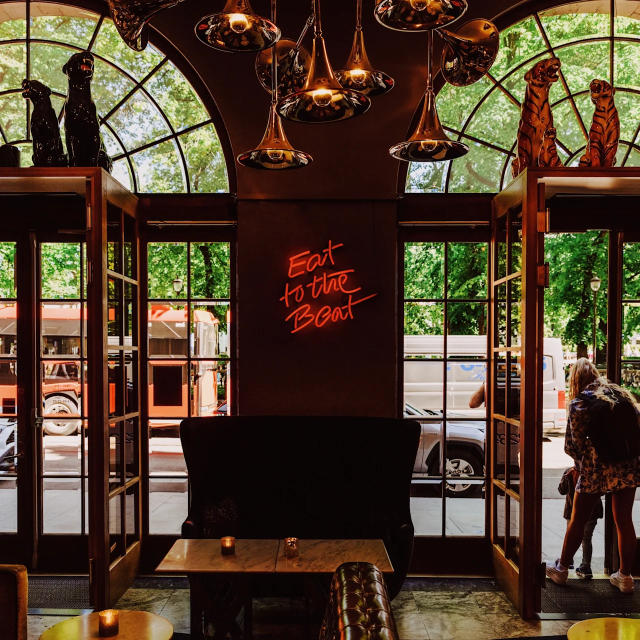 A cozy restaurant lounge features tufted seating, decorative lighting, and an inviting atmosphere. Neon text reads "Eat to the Beat" on a wall. Large windows show a red vehicle outside.