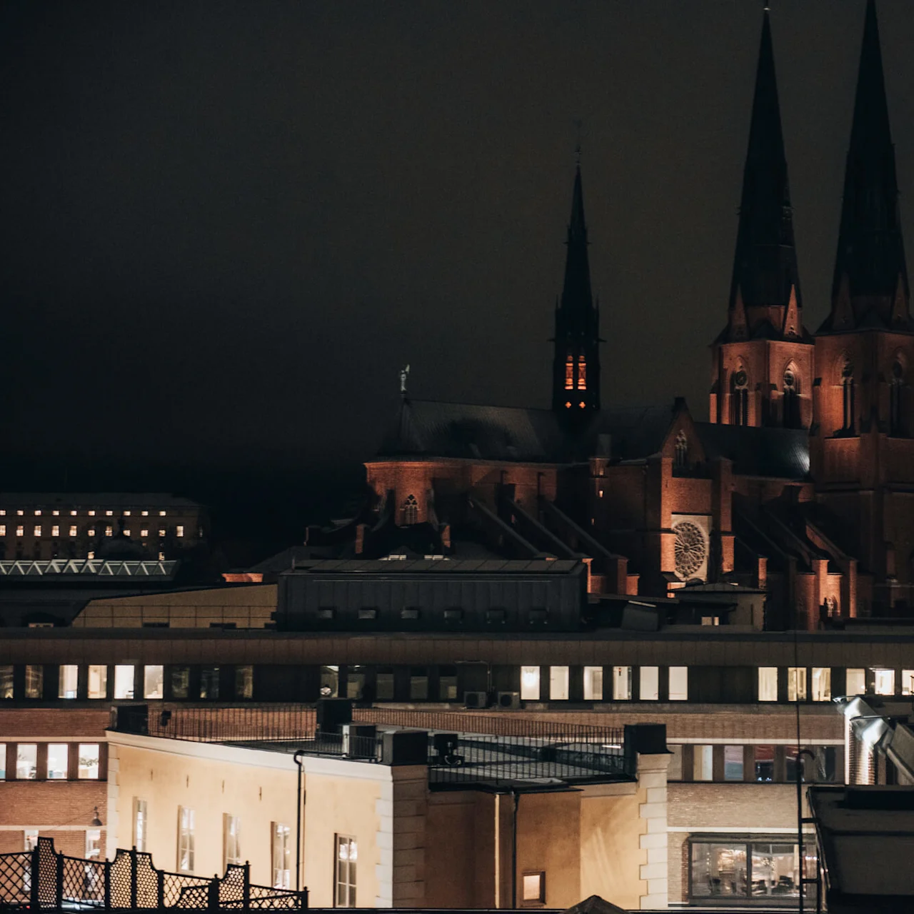 A large cathedral with illuminated spires stands against a dark night sky, surrounded by lower urban buildings with lit windows.