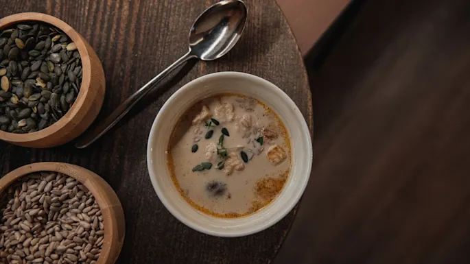 En varm bolle kremet suppe med urter og frø, ledsaget av skåler med gresskar- og solsikkefrø, på et rustikt trebord.
