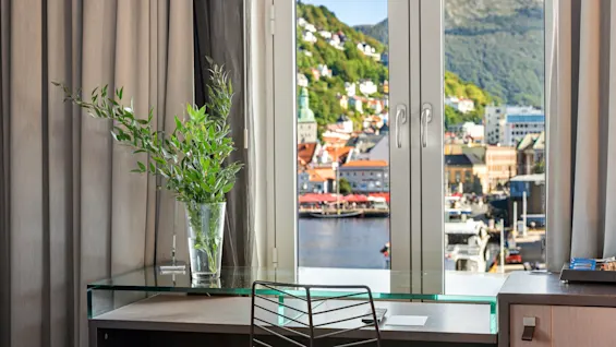 Vase med grønne blade står på et glasbord ved siden af en stol, nær et vindue med udsigt over en livlig by og bjerglandskab.