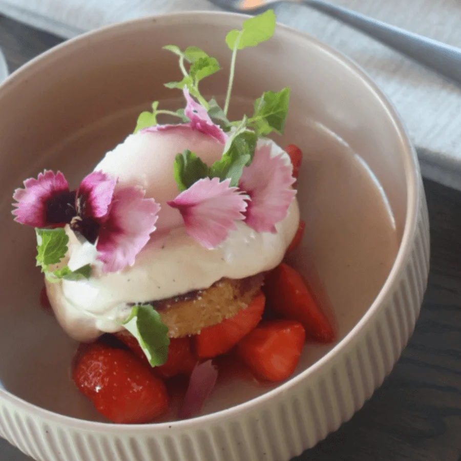 A dessert bowl holds a biscuit topped with cream, pink flowers, and microgreens, surrounded by sliced strawberries. Forks are placed on a table in the background.