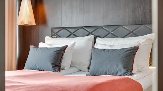 A bed with white linens and gray pillows is covered by a coral blanket in a modern room. A quilted headboard and pendant light complete the cozy setting.