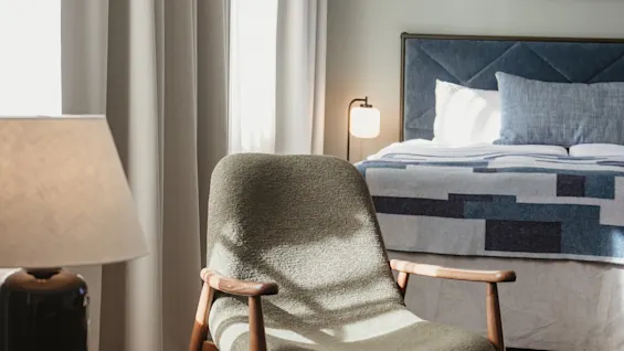 Ett mysigt hotellrum med en säng, en bekväm fåtölj badad i solljus och ett sidobord med en lampa. Home Hotel Post.