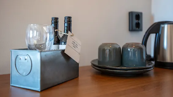 A metal container holds wine glasses and bottles, next to ceramic cups on a wooden surface.