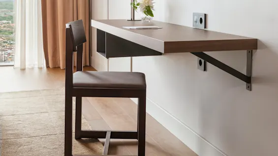 Ett skrivbord i trä har en lampa och en blomstervas, tillsammans med en stol. En stor TV är monterad på väggen ovanför. Solljus strömmar genom beige gardiner vid fönstret.