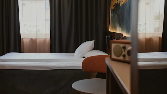 Et koselig hotellrom med en pent oppredd seng, mørke gardiner og en vintage radio på et nattbord. Teksten 'farg' er synlig på vinduet.