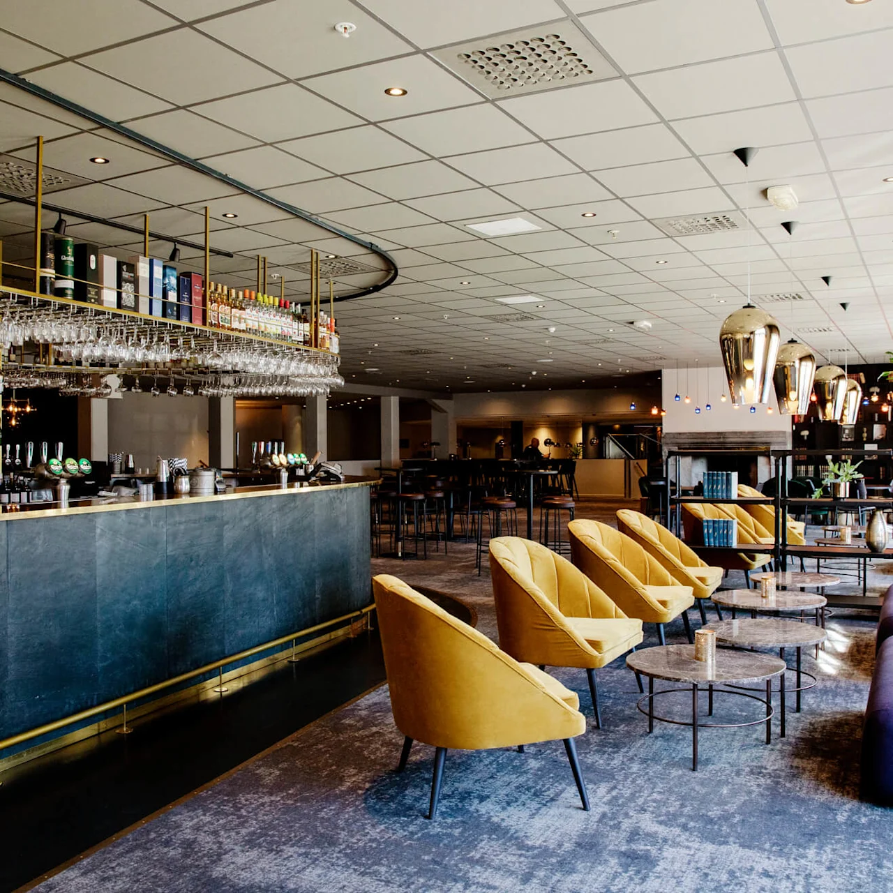 Bardisk fylld med flaskor och glas, bredvid mjuka gula stolar och små bord i en lounge med modern inredning och stora fönster som släpper in naturligt ljus.