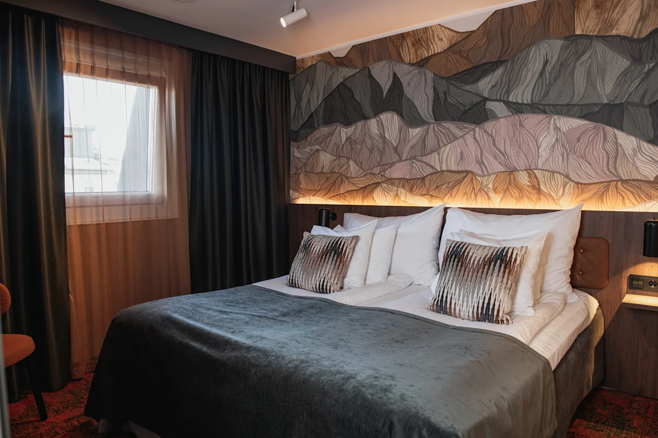 Et koselig hotellrom med en stor seng med grått teppe, et fjellinspirert veggmaleri og et vindu med gardiner.