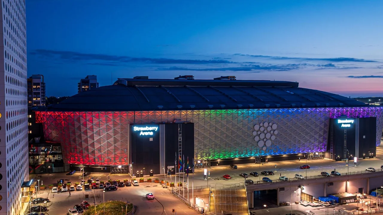 En stor arena viser fargerike lys over fasaden ved skumring, med parkerte biler i nærheten. Tekst på bygningen lyder "Strawberry Arena," under en klar kveldshimmel.