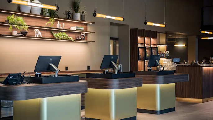 Kolme puista vastaanottotiskiä, joissa on tietokoneen näytöt, valaistu modernissa sisätilassa, jossa koristeelliset seinähyllyt sisältävät kasveja ja keramiikkaa. Kattovalaisimet parantavat tunnelmaa.