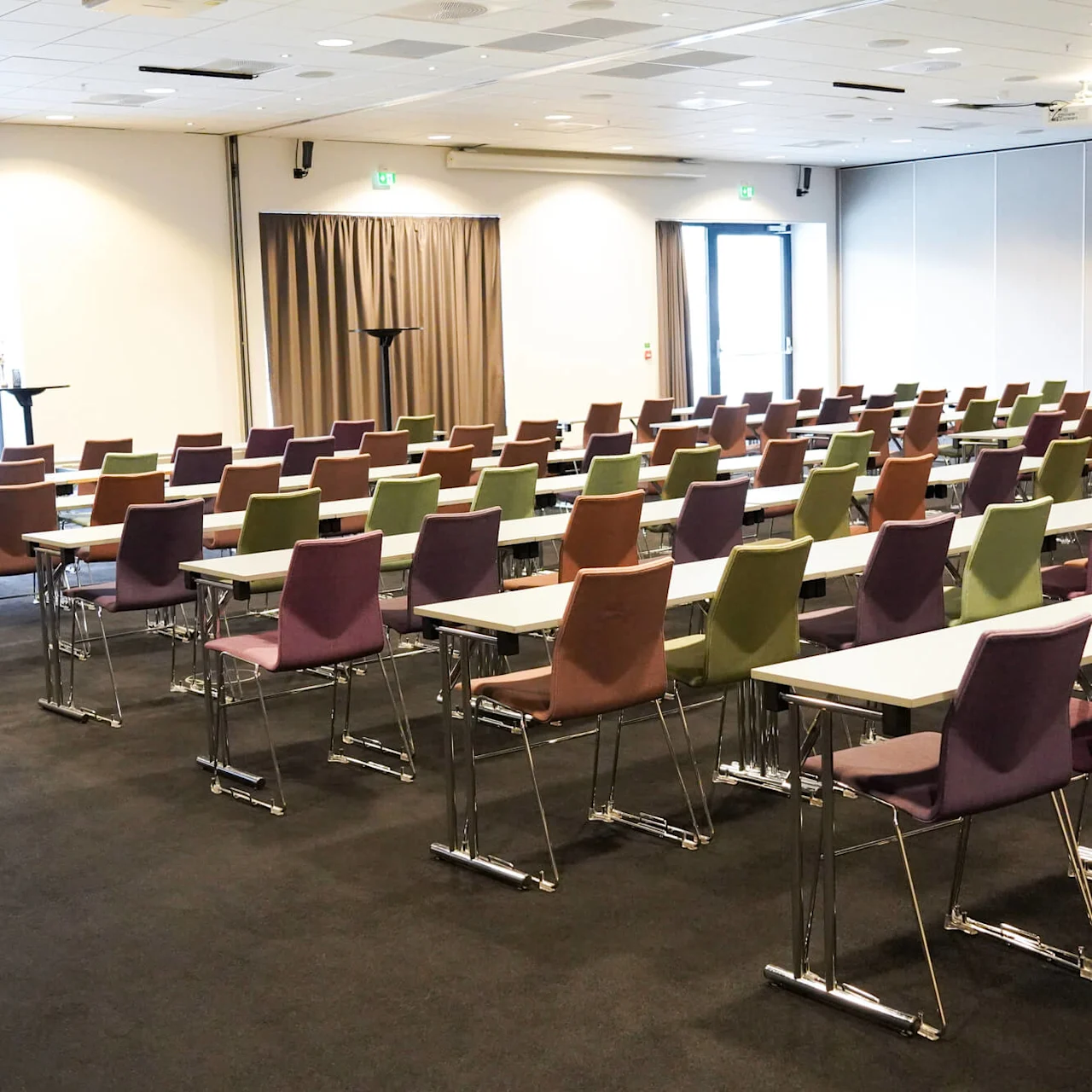 Rader med fargerike stoler plassert foran lange bord, arrangert i et romslig konferanserom. Store vinduer og nøytrale vegger skaper en lys, åpen atmosfære. Ingen tekst tilstede.