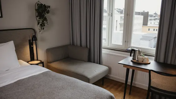 En pent redt seng med et grått teppe er ved siden av en divan. Nær ved står et lite bord med en vannkoker og kopper ved et lyst vindu med gardiner.