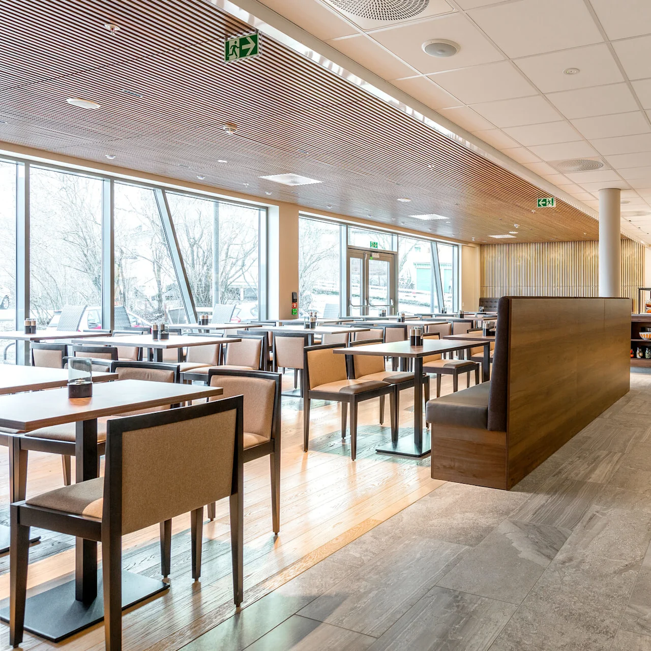 Moderni kahvilan sisätila, jossa pöytiä, tuoleja ja ruokailualuetta, valaistu luonnonvalolla suurista ikkunoista jotka näyttävät puiden ympäröimälle ulkoalueelle.