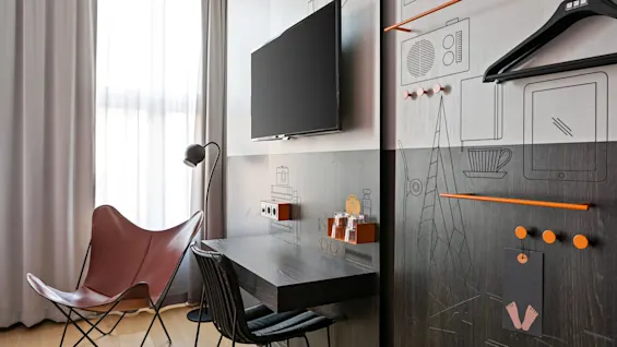 En brun skinnstol står ved siden av en veggmontert TV og pult under mykt lys i et moderne rom med illustrerte veggdesigner, inkludert kleshengere og hyller.