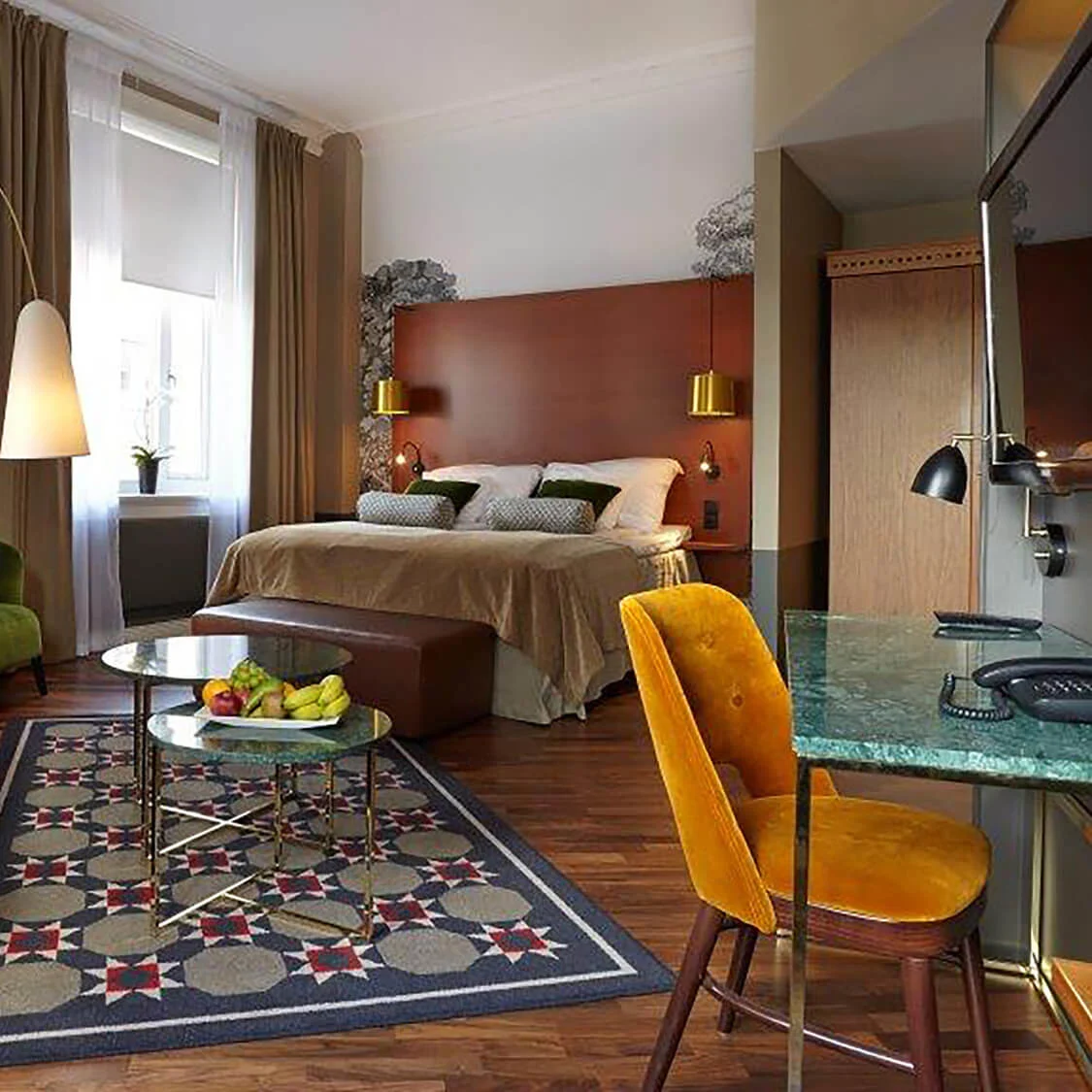 A well-lit hotel room with a bed, chairs, a desk, lamps, and a TV, reflecting a cozy and modern atmosphere.