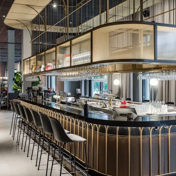 A curved bar counter with high chairs is set in a modern, spacious restaurant. Hanging wine glasses and ambient lighting add elegance, surrounded by lush plants and dining tables.