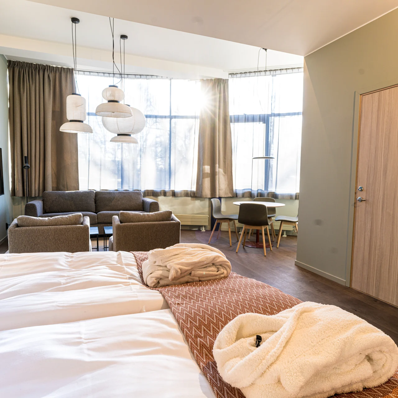 A neatly arranged hotel room features a bed with robes, a sitting area with armchairs and a sofa, a small dining table, and large windows allowing natural light.