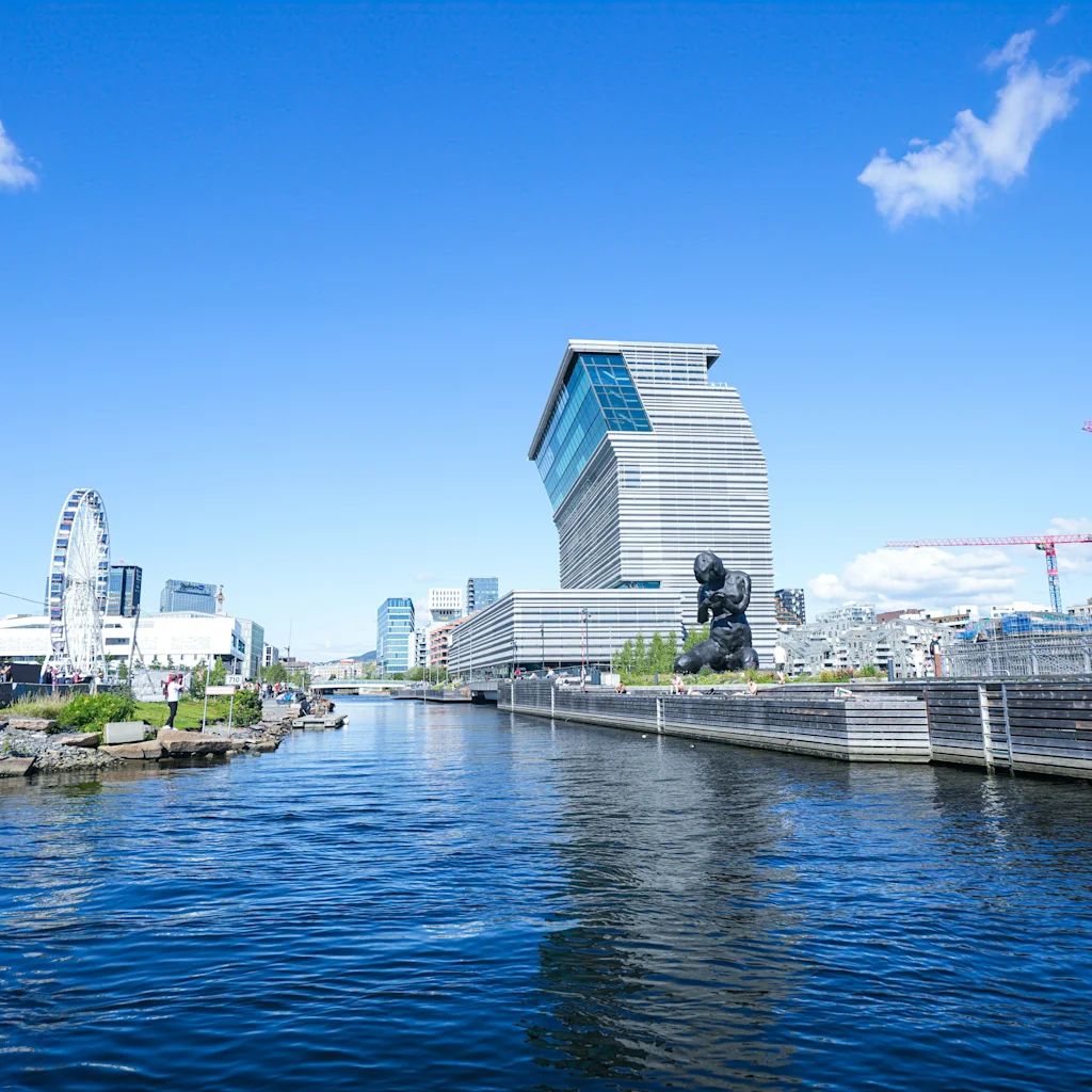 Et livlig bybilde ved vannet med moderne arkitektur, et pariserhjul og byggekraner under en klarblå himmel.