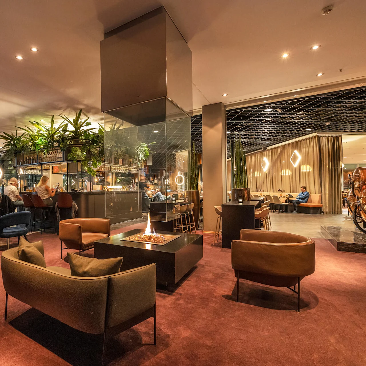 A cozy lounge features modern chairs surrounding a central fireplace, with patrons enjoying drinks at a bar. Green plants hang from above, creating a warm, inviting atmosphere.
