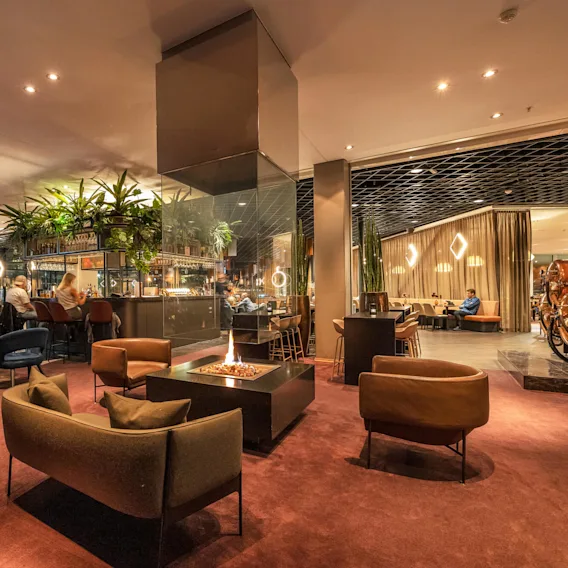En koselig lounge har moderne stoler rundt en sentral peis, med gjester som nyter drinker ved en bar. Grønne planter henger ovenfra, og skaper en varm, innbydende atmosfære.