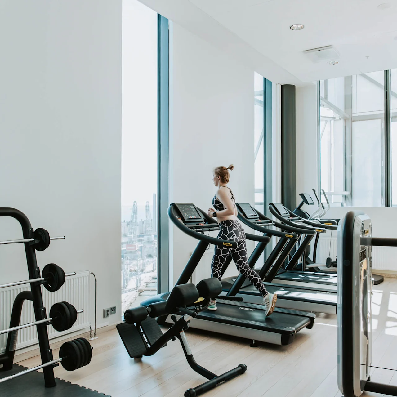 En person løber på et løbebånd i et lyst, rummeligt fitnesscenter med store vinduer og udsigt over en by. Tæt på er der vægtløfteudstyr.